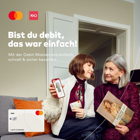 Zwei Frauen sitzen auf einer Couch und lächeln sich an. Die eine hält ein Smartphone, auf dem eine Debitkarte abgebildet ist, die andere ein Modemagazin in der Hand. Der Text auf Deutsch wirbt für einfaches und sicheres Bezahlen mit einer Debit Mastercard.