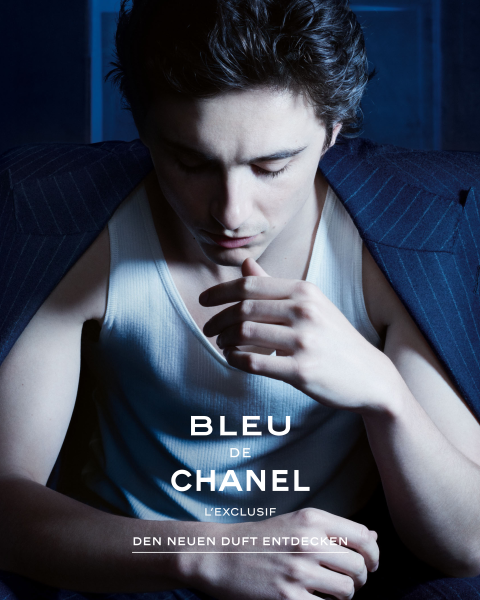 Ein junger Mann in einem Nadelstreifenanzug und einem weißen Tank-Top sitzt mit gesenktem Kopf und einer Hand an den Lippen. Das Bild zeigt den Text "BLEU DE CHANEL L'EXCLUSIF DEN NEUEN DUFT ENTDECKEN", der für einen Chanel-Duft wirbt.