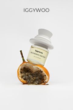 Ein Parfümflakon mit der Aufschrift "Bohemian Water" von IGGYWOO steht auf einer halbierten Passionsfrucht mit freiliegenden Kernen vor einem schlichten weißen Hintergrund.