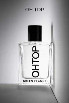 Ein durchsichtiger Parfümflakon mit der Aufschrift "OHTOP", einem schwarzen Verschluss und der Aufschrift "GREEN FLANNEL" am Boden, der an einer weißen Wand lehnt, über der "OH TOP" steht.