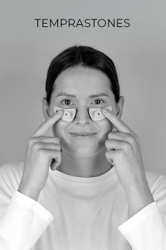 Eine Person in einem weißen Hemd lächelt, während sie zwei kleine, gebogene Objekte unter ihren Augen hält. Über ihrem Kopf erscheint das Wort "TEMPRASTONES".