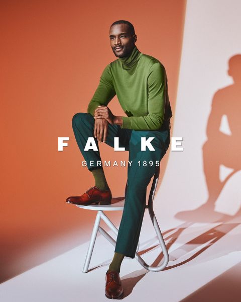 Ein Mann in einem grünen Rollkragenpullover, einer dunkelgrünen Hose und braunen Schuhen sitzt auf einem weißen Hocker vor einem orangefarbenen und weißen Hintergrund. Der Text "FALKE GERMANY 1895" ist über das Bild gelegt.
