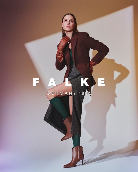 Ein Modell in einem kastanienbraunen Blazer, braunen Handschuhen, einer grünen Strumpfhose, einem grauen Rock mit hohen Schlitzen und braunen Ankle Boots posiert vor einem geometrischen Hintergrund. Der Text lautet "FALKE GERMANY 1895".