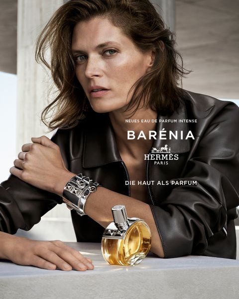 Eine Frau in Lederjacke posiert mit einem Flakon des Parfums Hermès Barénia und stützt ihren Arm auf eine Fläche. Der Text auf dem Bild lautet: "NEUES EAU DE PARFUM INTENSE BARÉNIA HERMÈS PARIS DIE HAUT ALS PARFUM.