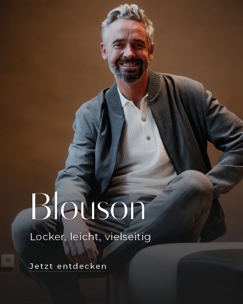 Ein lächelnder Mann mit grauem Haar und Bart sitzt lässig mit einem gekreuzten Bein da. Er trägt eine graue Blousonjacke über einem hellen Hemd. Der Text auf Deutsch lautet: "Blouson. Locker, leicht, vielseitig. Jetzt entdecken.