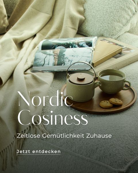 Eine gemütliche Szene mit einem beigen Sofa, einer aufgeschlagenen Zeitschrift und einem Holztablett mit einer Teekanne, einer Tasse und drei Keksen. Der Text lautet: "Nordische Gemütlichkeit. Zeitlose Gemütlichkeit Zuhause. Jetzt entdecken.