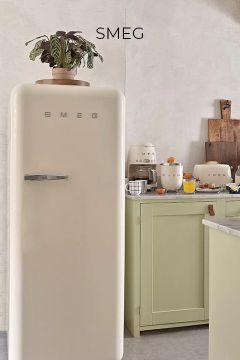 Ein cremefarbener SMEG-Kühlschrank steht in einer hellen Küche mit hellgrünen Schränken, einer Topfpflanze auf dem Dach und verschiedenen kleinen Küchengeräten und Frühstücksartikeln auf dem Tresen.