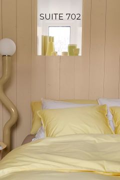 Ein ordentlich gemachtes Bett mit gelbem Bettzeug steht neben einer geschwungenen Stehlampe. Über dem Bett befindet sich ein kleines Fenster mit dekorativen Gegenständen und an der Wand steht "SUITE 702".