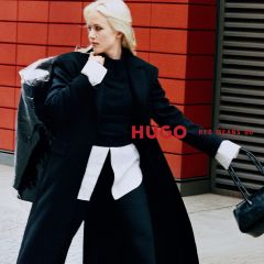 Eine Frau in einem schwarzen Mantel und einem weißen Hemd posiert neben dem Schriftzug „HUGO RED MEANS GO“.