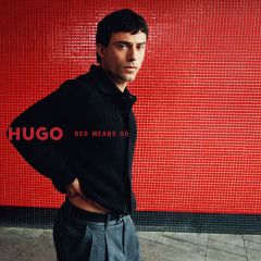 Ein Mann in einer schwarzen Strickjacke und einer grauen Hose steht vor einer Wand aus roten Fliesen, auf der der Schriftzug „HUGO RED MEANS GO“ zu lesen ist.