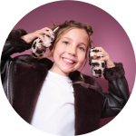 Lächelndes junges Mädchen, das eine braune Kunstpelzjacke und Ohrenschützer mit Tiermotiven trägt, die Ohrenschützer an die Ohren hält und vor einem rosa Hintergrund steht.