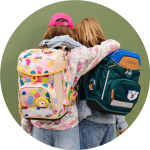 Zwei Kinder mit bunten Rucksäcken, die ihre Arme um die Schultern des anderen legen. Das eine hat einen beigen Rucksack mit bunten Flecken, das andere einen dunkelgrünen Rucksack mit Tierflecken. Grüner Hintergrund.