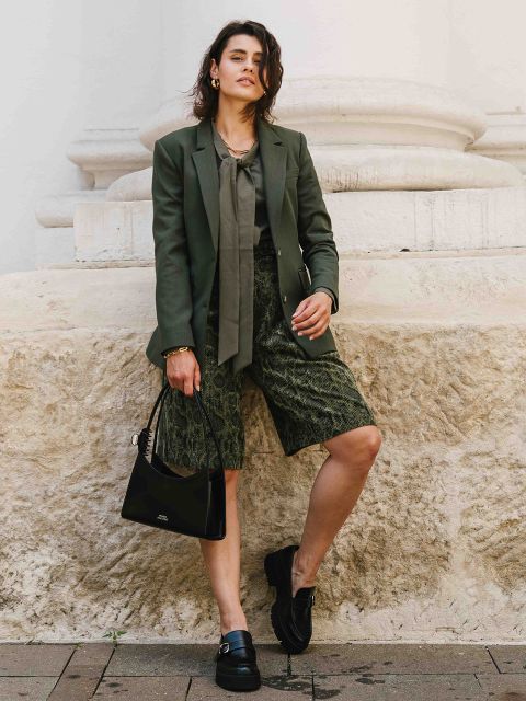 Eine Frau in einem grünen Blazer, einer passenden Bluse mit Krawattenausschnitt und gemusterten grünen knielangen Shorts steht vor einer hellen Steinmauer, hält eine schwarze Handtasche in der Hand und trägt schwarze Slipper.