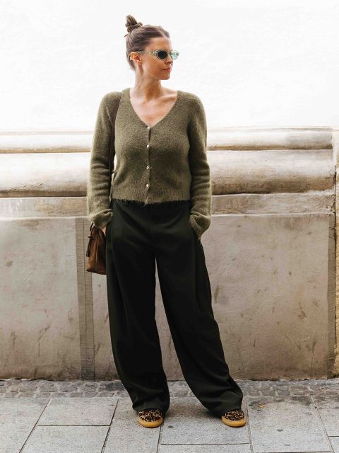 Eine Frau mit Haarknoten trägt eine Sonnenbrille, eine grüne Strickjacke mit Knöpfen, eine weite schwarze Hose, Schuhe mit Leopardenmuster und eine braune Umhängetasche, während sie an einer Steinmauer steht.