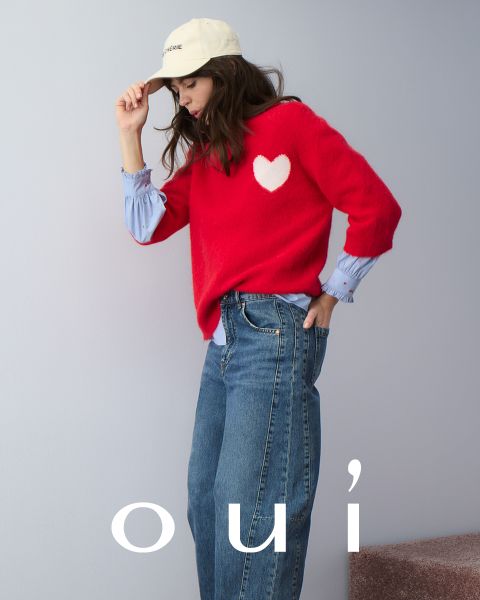 Eine Frau, die einen roten Pullover mit einem weißen Herz, blaue Jeans und eine weiße Mütze trägt, posiert vor einer hellen Wand. Am unteren Rand des Bildes steht in weißen Buchstaben das Wort "oui".