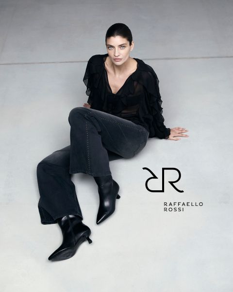 Eine Frau mit dunklem Haar sitzt auf einem hellgrauen Boden und trägt eine schwarze Rüschenbluse, dunkle Jeans und schwarze spitze Stiefel. Neben ihr ist das Logo von Raffaello Rossi zu sehen.