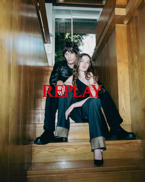 Ein Mann und eine Frau sitzen in dunkler, stilvoller Kleidung auf einer Holztreppe in einem Innenraum. Das Wort "REPLAY" ist in der Mitte des Bildes in roten Großbuchstaben prominent eingeblendet.