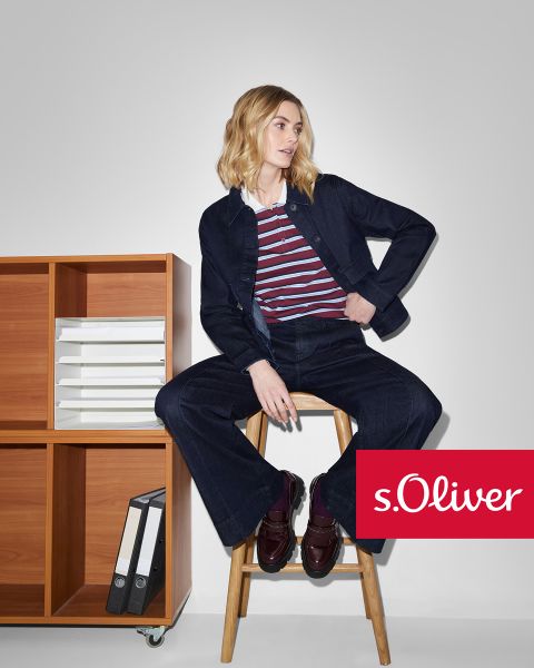 Eine Frau in dunkler Jeansjacke und Hose, die ein gestreiftes Oberteil trägt, sitzt auf einem Holzschemel neben einem Holzregal mit Akten. Das s.Oliver-Logo erscheint in der unteren rechten Ecke.