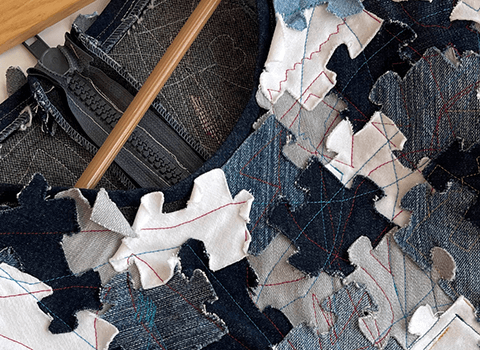 Nahaufnahme eines Kleidungsstücks auf einem Kleiderbügel mit sich überlappenden blattförmigen Aufnähern aus Denim und weißem Stoff mit sichtbaren Nähten in verschiedenen Farben und einem sichtbaren Reißverschluss auf der Rückseite.