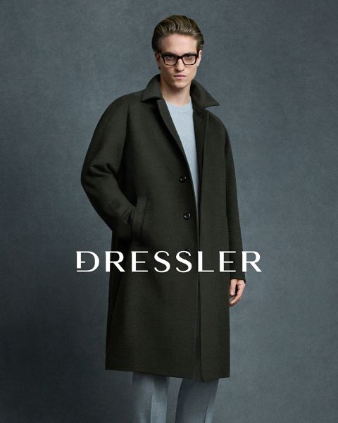 Ein Mann, der eine Brille, einen dunklen Mantel und eine hellgraue Hose trägt, steht vor einem grauen Hintergrund. Das Wort "DRESSLER" ist in großen weißen Buchstaben quer über das Bild gedruckt.