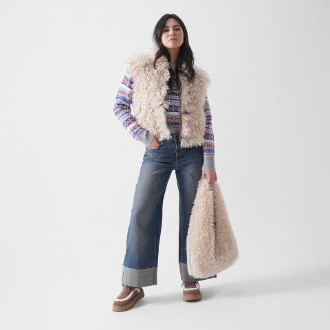 Eine Frau posiert vor einem schlichten weißen Hintergrund und trägt eine flauschige beigefarbene Weste, einen bunt gemusterten Pullover, eine Jeans mit weitem Bein, hellbraune Plateauschuhe und eine passende beigefarbene Pelztasche.