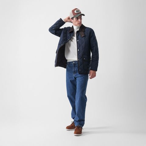 Eine Person, die eine marineblaue Steppjacke, einen weißen Rollkragenpullover, blaue Jeans, braune Bootsschuhe und eine braun-gelbe Baseballmütze trägt, steht in entspannter Pose vor einem schlichten weißen Hintergrund.