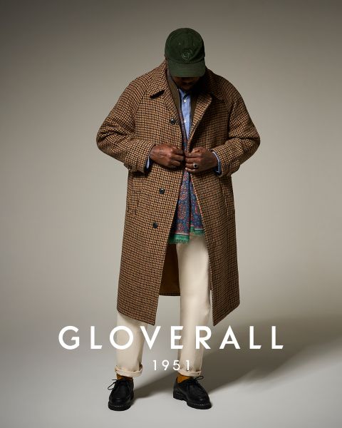Eine Person, die einen karierten Mantel, eine grüne Mütze, ein blaues Hemd, einen gemusterten Schal, eine cremefarbene Hose und schwarze Schuhe trägt, blickt nach unten. Unten auf dem Bild steht der Text "Gloverall 1951".