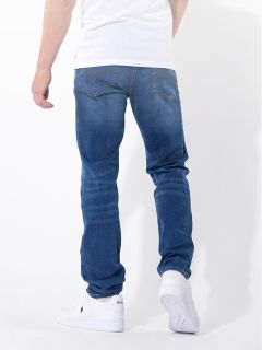 Eine Person, die blaue Jeans, ein weißes T-Shirt und weiße Turnschuhe trägt, steht mit dem Rücken zur Kamera vor einem schlichten weißen Hintergrund.