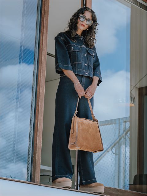 Eine Frau, die eine dunkle Jeansjacke und weite Hosen trägt, steht auf einem Balkon und hält eine hellbraune Handtasche in der Hand. Sie hat gewelltes Haar, trägt eine Sonnenbrille und Plateauschuhe. Im Hintergrund sind der blaue Himmel und das Glasgeländer zu sehen.