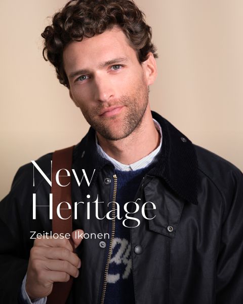 Ein Mann mit lockigen braunen Haaren und einem kurzen Bart, der eine dunkle Jacke, einen blauen Pullover und ein Hemd mit Kragen trägt, steht vor einem beigen Hintergrund. Der Text auf dem Bild lautet "New Heritage" und "Zeitlose Ikonen".