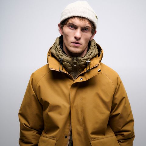 Ein junger Mann, der einen senfgelben Wintermantel, einen olivgrünen Kapuzenpulli und eine cremefarbene Strickmütze trägt, steht vor einem schlichten, hellen Hintergrund und schaut mit neutralem Blick direkt in die Kamera.