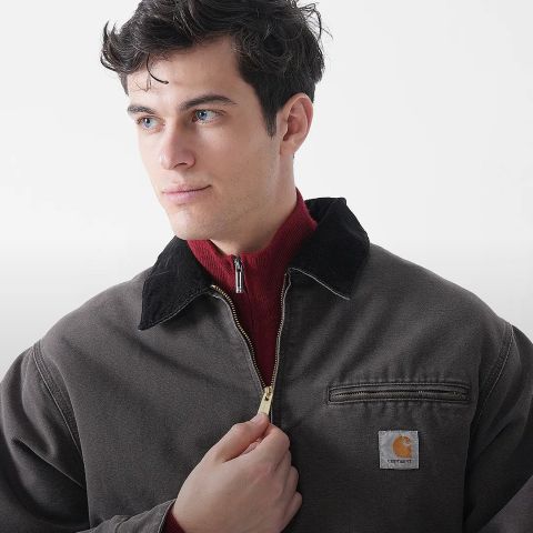 Ein junger Mann mit kurzen dunklen Haaren trägt eine braune Jacke mit einem schwarzen Kragen über einem roten Pullover mit Reißverschluss. Er blickt zur Seite und öffnet teilweise den Reißverschluss seiner Jacke. Das Carhartt-Logo ist auf der Jacke zu sehen.