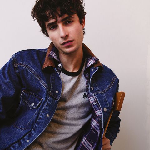 Ein junger Mann mit lockigen dunklen Haaren trägt eine Jeansjacke über einem karierten Hemd und einem grauen T-Shirt, lehnt sich leicht vor und blickt in die Kamera vor einem schlichten hellen Hintergrund.