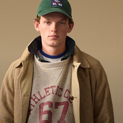 Ein junger Mann, der eine grüne Baseballmütze, einen hellbraunen Fischgrätenmantel, ein kariertes und ein graues Sweatshirt mit dem Aufdruck "ATHLETIC C 67" auf der Vorderseite trägt, steht vor einem beigen Hintergrund und schaut in die Kamera.