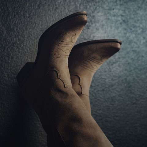 Nahaufnahme eines Paars brauner Cowboystiefel aus Wildleder mit Ziernähten vor einem dunklen, strukturierten Hintergrund. Die Stiefel sind nach oben gerichtet, was ihr Design und ihr Material hervorhebt.