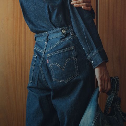 Eine Person, die ein dunkles Jeanshemd und Jeans trägt, steht vor einer Holzwand und hält in einer Hand eine Jeansjacke, während sie der Kamera den Rücken zuwendet.