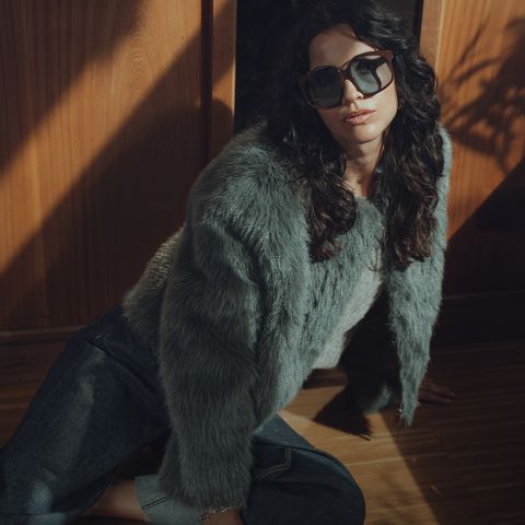 Eine Frau mit gewelltem dunklem Haar, die eine übergroße Sonnenbrille, eine graue Pelzjacke und dunkle Jeans trägt, sitzt bei dramatischer Beleuchtung auf einem Holzboden in der Nähe einer Holzwand.