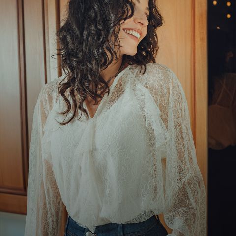 Eine Frau mit gewelltem dunklem Haar, die eine weiße Spitzenbluse und blaue Jeans trägt, steht lächelnd in einem warm beleuchteten, holzgetäfelten Raum und schaut zur Seite.