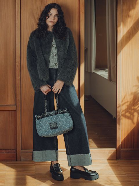 Eine Frau mit gewelltem dunklem Haar trägt einen grauen Pelzmantel, einen grauen Pullover, Jeans mit weitem Bein und schwarze Plateauschuhe und hält eine blaue, gesteppte Handtasche in der Hand, während sie in einem sonnenbeschienenen, holzgetäfelten Raum steht.