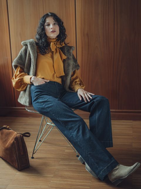 Eine Frau mit gewelltem dunklem Haar sitzt auf einem Stuhl und trägt eine senfgelbe Bluse mit Schleife, eine Jeans mit weitem Bein, eine Weste aus Kunstpelz und hellbraune Stiefel, während neben ihr eine braune Wildledertasche auf dem Boden liegt. Sie posiert selbstbewusst in einem holzgetäfelten Raum.