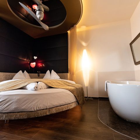 Ein modernes Hotelzimmer mit einem runden Bett, beigem Bettzeug, einer weißen Stehlampe, einem gekippten Wandspiegel und einer großen weißen Badewanne auf dunklem Holzfußboden bei gedämpfter Beleuchtung.