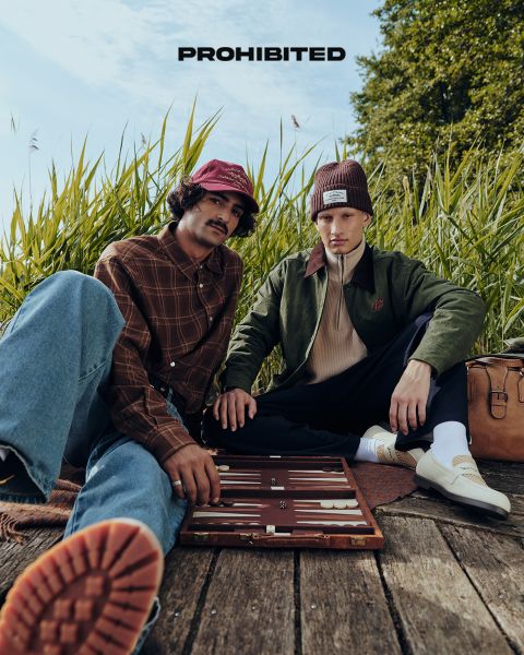 Zwei Männer sitzen auf einem Holzdeck im hohen Gras und spielen Backgammon. Der eine trägt eine rote Mütze, ein kariertes Hemd und Jeans, der andere eine kastanienbraune Mütze, eine olivfarbene Jacke und eine dunkle Hose. Über ihnen steht "PROHIBITED" geschrieben.