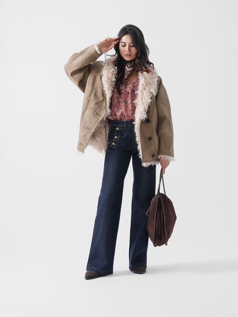 Eine Frau posiert in einer hellbraunen Shearling-Jacke über einem gemusterten Hemd, hochtaillierten Schlagjeans und braunen Stiefeln und hält eine große braune Tasche in ihrer linken Hand vor einem schlichten weißen Hintergrund.