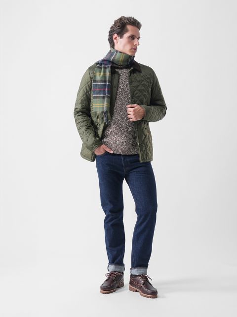 Eine Person steht mit dem Gesicht nach links und trägt eine grüne Steppjacke, einen karierten Schal, einen grauen Pullover, blaue Jeans und braune Bootsschuhe vor einem schlichten weißen Hintergrund.