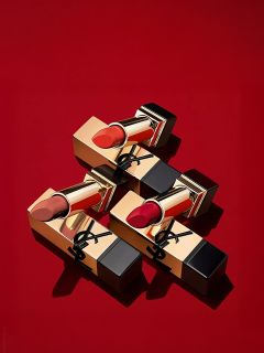 Drei goldene und schwarze Lippenstifte mit verschiedenen Rottönen, die diagonal auf einem einfarbig roten Hintergrund angeordnet sind. Die Verpackung ist mit einem schwarzen Logo auf der goldenen Hülle versehen.