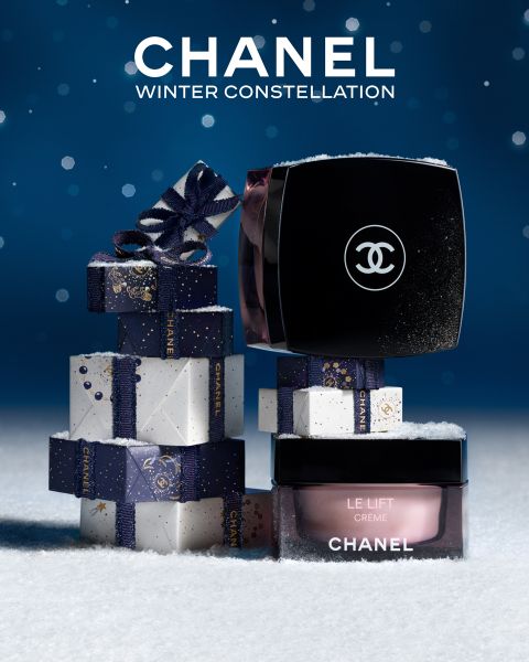Die Cremetiegel von Chanel Le Lift stehen auf einem verschneiten Boden mit gestapelten Chanel-Geschenkboxen mit Schleifenband. Der Hintergrund ist blau mit weißen Sprenkeln, und der Text lautet "CHANEL WINTER CONSTELLATION".