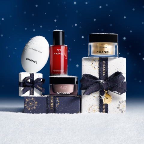 Chanel-Pflegeprodukte und -Geschenkschachteln werden auf Schnee mit dunkelblauem, sternförmigem Hintergrund präsentiert. Schnee bedeckt die Produkte und Schachteln, die mit dunklen Schleifen und goldenen Akzenten verziert sind.