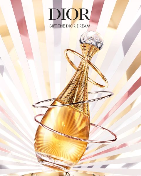 Ein goldener Dior-Parfümflakon mit spiralförmigen Ringen vor einem strahlenden Hintergrund mit Sonnenaufgang. Der Text darüber lautet: "DIOR" und "GIFT THE DIOR DREAM".