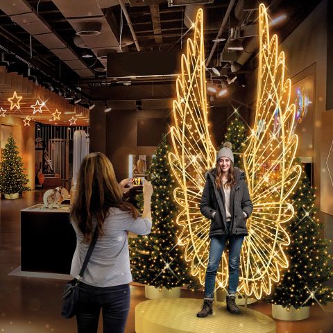 Eine Frau macht ein Foto von einer anderen Frau, die vor großen, leuchtenden Engelsflügeln aus Lichtern in einer festlich geschmückten Halle mit Weihnachtsbäumen steht.