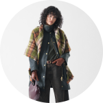 Eine Person mit lockigem Haar, die einen grünen Mantel, einen karierten Schal, braune Handschuhe und eine dunkle Hose trägt, hält eine glänzende lila Handtasche und steht vor einem schlichten weißen Hintergrund.